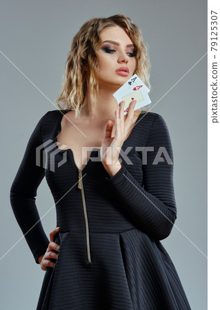 Blonde female in black dress showing two playing cards, posing against gray background. Gambling entertainment, poker, casino. Close-up. 79125307