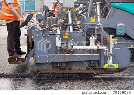 A road worker operates an asphalt paver and pours asphalt concrete while standing behind a control panel on a metal step on a summer day. A road worker operates an asphalt paver and pours asphalt concrete while standing behind a control panel on a metal step on a summer day. 79129089