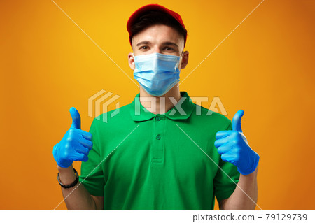 Young delivery man in green t-shirt and cap wearing face mask against yellow background Young delivery man in green t-shirt and cap wearing face mask against yellow background 79129739