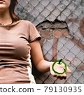 A young caucasian cool urban girl holding a draw green apple in front of a rough shabby concrete and brick wall with graffiti and a steel netting fence 79130935