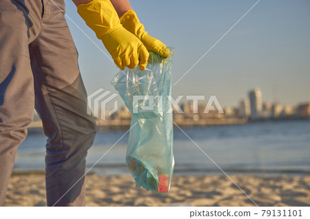 Young volunteer in yellow gloves is walking with garbage bag along a dirty beach of the river and cleaning up trash. People and ecology. Close-up. 79131101