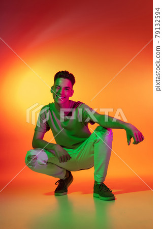Portrait of Caucasian young man sitting isolated on orange studio background in neon light. Concept of human emotions, facial expression. Portrait of Caucasian young man sitting isolated on orange studio background in neon light. Concept of human emotions, facial expression. 79132594