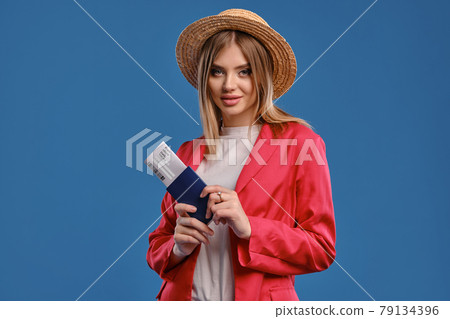 Blonde female in straw hat, white blouse and red pantsuit. She smiling, holding passport and ticket while posing on blue studio background. Close-up 79134396