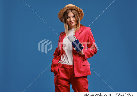 Blonde model in straw hat, white blouse and red pantsuit. She smiling, showing passport and ticket while posing on blue studio background. Close-up Blonde model in straw hat, white blouse and red pantsuit. She smiling, showing passport and ticket while posing on blue studio background. Close-up 79134463