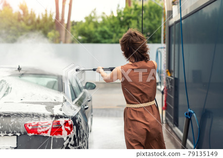 Back view young adult woman washing car with high pressure water equipment pump self-service outdoor on bright windy summer day. Vehicle covered foam shampoo chemical detergents carwash self service 79135149
