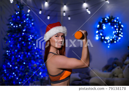 Beautiful red-haired female athlete in a Santa Claus hat posing in a dark studio. Fitness woman in sportswear holds a dumbbell in her hands on the background of the New Year tree. Beautiful red-haired female athlete in a Santa Claus hat posing in a dark studio. Fitness woman in sportswear holds a dumbbell in her hands on the background of the New Year tree. 79138178
