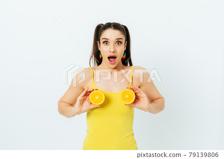 Young brunette woman wearing summer outfit holding halves of oranges near her chest 79139806