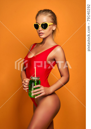 Lovely young female in swimwear and sunglasses holding glass of fresh drink while standing on yellow, orange hot background 79140292
