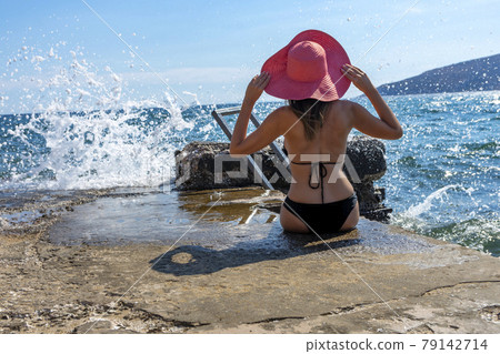 Tanning woman wearing red sun hat sitting on a concrete pier while waves splashing in front of her - rear view Tanning woman wearing red sun hat sitting on a concrete pier while waves splashing in front of her - rear view 79142714