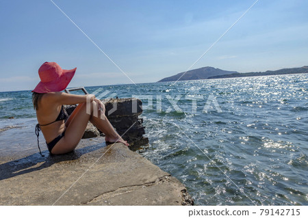 Tanning woman wearing red sun hat sitting on a concrete pier side view 79142715