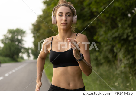 Young female runner, athlete is jogging at road in summer sunshine. Beautiful caucasian woman training, listening to music. Concept of sport, healthy lifestyle, movement, activity. Young female runner, athlete is jogging at road in summer sunshine. Beautiful caucasian woman training, listening to music. Concept of sport, healthy lifestyle, movement, activity. 79146450