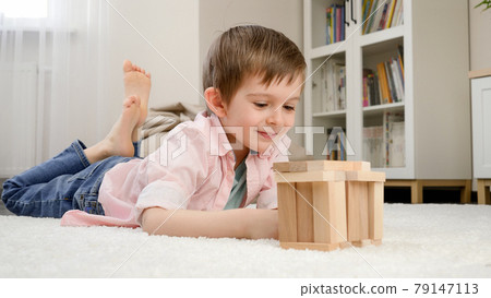 Cute smiling boy lying on floor and looking at toy house he built from toy wooden blocks. Concept of child education and games at home Cute smiling boy lying on floor and looking at toy house he built from toy wooden blocks. Concept of child education and games at home 79147113
