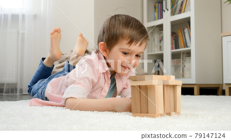 Smiling boy lying on floor and building house or tower from wooden blocks. Concept of child education and games at home Smiling boy lying on floor and building house or tower from wooden blocks. Concept of child education and games at home 79147124