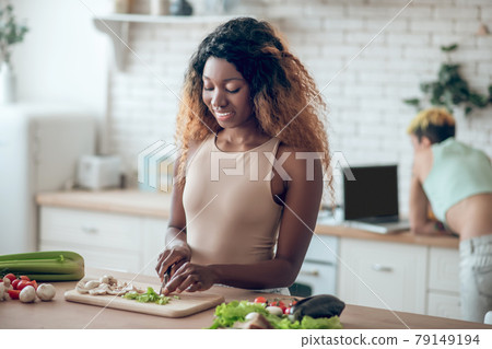 Dark-skinned woman chopping vegetables and girlfriend at distance Dark-skinned woman chopping vegetables and girlfriend at distance 79149194