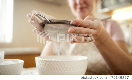 Closeup of young woman sifting flour through metal sieve on domestic kitchen Closeup of young woman sifting flour through metal sieve on domestic kitchen 79149257