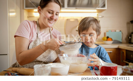 Young smiling mother cooking with little son and sifting flour with sieve. Children cooking with parents, little chef, family having time together, domestic kitchen. Young smiling mother cooking with little son and sifting flour with sieve. Children cooking with parents, little chef, family having time together, domestic kitchen. 79149311