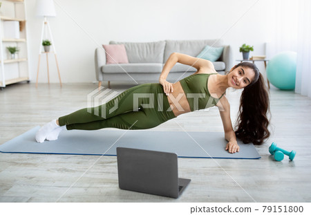Online sports during covid. Fit Indian woman doing exercises in front of laptop, standing in side plank at home Online sports during covid. Fit Indian woman doing exercises in front of laptop, standing in side plank at home 79151800