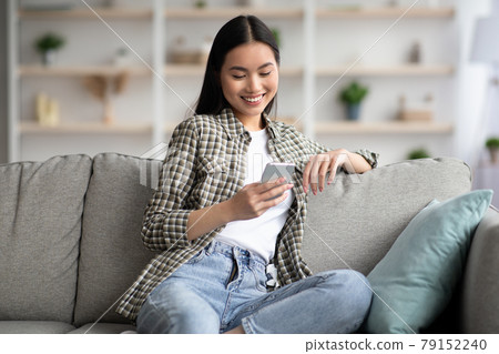 Cheerful asian lady sitting on sofa and using smartphone Cheerful asian lady sitting on sofa and using smartphone 79152240
