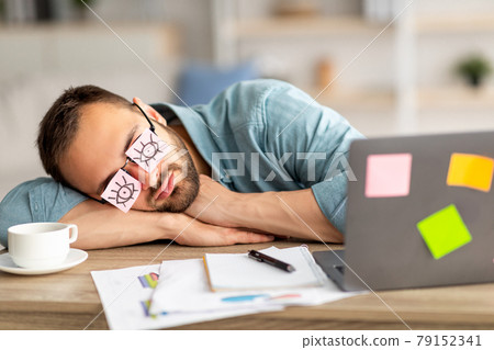 Lazy unproductive young guy wearing funny sticky notes with open eyes on his glasses, sleeping at workplace 79152341