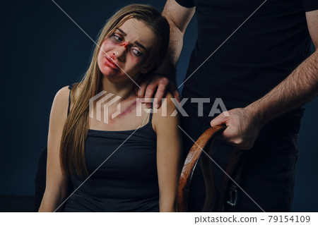 Cruel man put his hand on female shoulder, showing belt. Victim with bruises on face sitting nearby. Blue background. Domestic violence. Close-up. Cruel man put his hand on female shoulder, showing belt. Victim with bruises on face sitting nearby. Blue background. Domestic violence. Close-up. 79154109