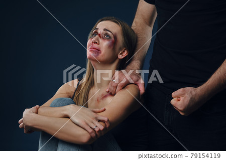 Strong man put his hand on female shoulder, showing fist. Victim with bruises on face sitting nearby. Blue background. Domestic violence. Close-up. 79154119