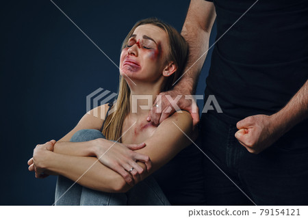 Strong man put his hand on female shoulder, showing fist. Victim with bruises on face sitting nearby. Blue background. Domestic violence. Close-up. 79154121