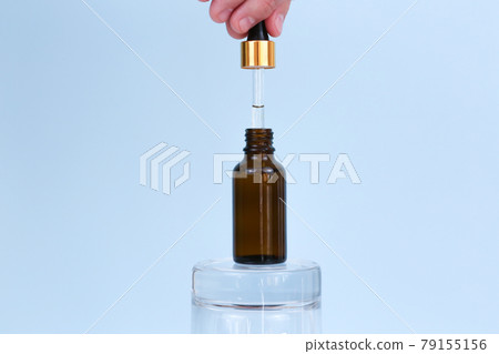 Male hand holds dropper with oil. Opened jar of natural cosmetic product on glass podium on blue background. Essential oil for care of skin. Skincare, moisturizing beauty product. Cosmetic production. 79155156
