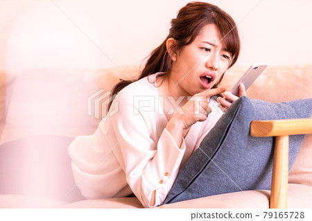 A moody woman lying on the sofa in the living room and looking at the screen of her smartphone 79165728