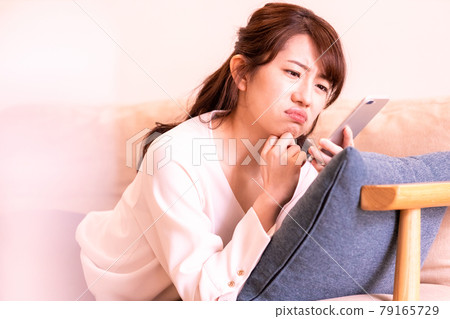 A moody woman lying on the sofa in the living room and looking at the screen of her smartphone 79165729