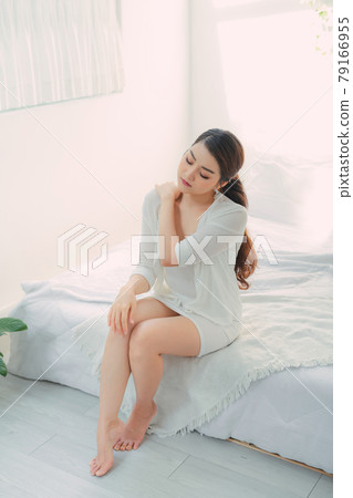 Beautiful girl sitting on bed, touches neck, smiling and keeps eyes closed Beautiful girl sitting on bed, touches neck, smiling and keeps eyes closed 79166955