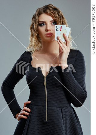 Blonde woman in black dress showing two playing cards, posing against gray background. Gambling entertainment, poker, casino. Close-up. 79173508