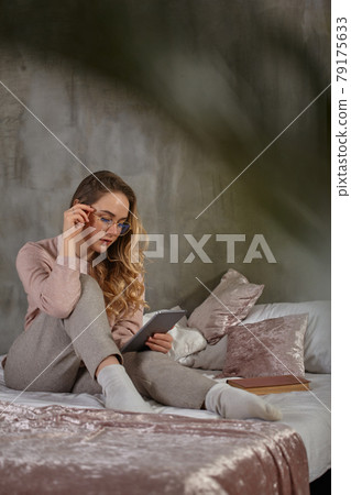 Blonde lady in glasses, casual clothing. Sitting on bed, working with a tablet in bedroom. Student, blogger. Interior with gray wall 79175633