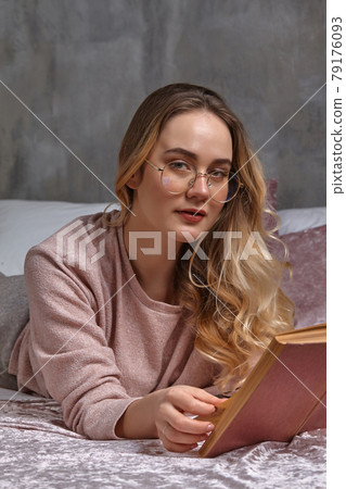 Blonde lady in glasses, casual clothing. She is looking at you, holding book, laying on bed in bedroom. Student, blogger, document studying. Close-up Blonde lady in glasses, casual clothing. She is looking at you, holding book, laying on bed in bedroom. Student, blogger, document studying. Close-up 79176093