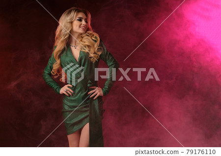 Gorgeous blonde curly lady in green stylish dress and jewelry. She has put her hands on waist, smiling, posing on colorful smoky background. Close up Gorgeous blonde curly lady in green stylish dress and jewelry. She has put her hands on waist, smiling, posing on colorful smoky background. Close up 79176110