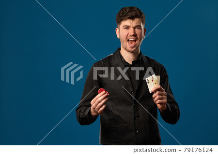 Newbie in poker, in black vest and white shirt. Holding two red chips and aces. Posing against blue background. Gambling, casino. Close-up. 79176124