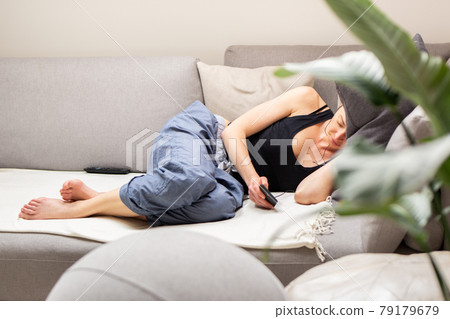Tired young woman, resting, lying on a comfortable sofa with big cushions in home living room using her mobile phone. Tired young woman, resting, lying on a comfortable sofa with big cushions in home living room using her mobile phone. 79179679