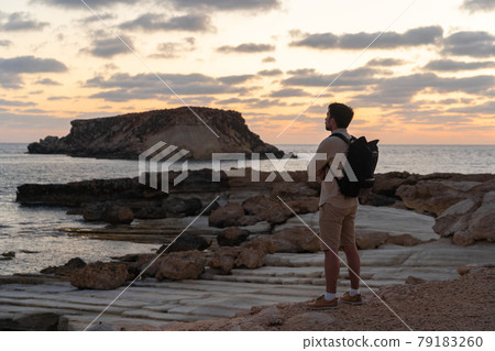 Man with backpack is hiking along a rocky coast at sunset near the Mediterranean sea in Cyprus. Male tourist admires the view Yeronisos, or Holy Island, lies off the coast of Agios Georgios Pegeias Man with backpack is hiking along a rocky coast at sunset near the Mediterranean sea in Cyprus. Male tourist admires the view Yeronisos, or Holy Island, lies off the coast of Agios Georgios Pegeias 79183260