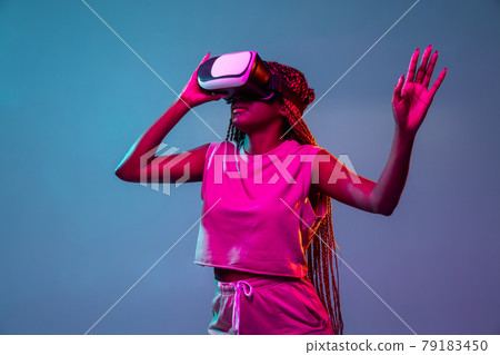 Portrait of young african woman in vr glasses on blue neon background. Concept of virtual reality, entertainment and people. Portrait of young african woman in vr glasses on blue neon background. Concept of virtual reality, entertainment and people. 79183450