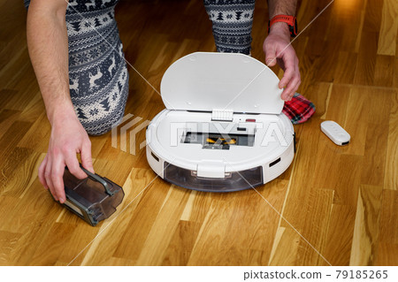 Caucasian young man in christmas pajamas doing vacuum cleaner robot maintenance. Male in winter patterned sleepwear and red socks slippers starts automatic vacuum cleaner. Robot vacuum cleaner repair Caucasian young man in christmas pajamas doing vacuum cleaner robot maintenance. Male in winter patterned sleepwear and red socks slippers starts automatic vacuum cleaner. Robot vacuum cleaner repair 79185265