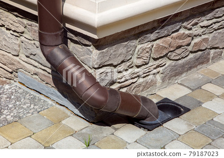 gutter made of brown pvc plastic drainage of rainwater from the roof of the house into the ground drainage system on the facade of the building lined with natural stone with a sidewalk made of tiles. gutter made of brown pvc plastic drainage of rainwater from the roof of the house into the ground drainage system on the facade of the building lined with natural stone with a sidewalk made of tiles. 79187023