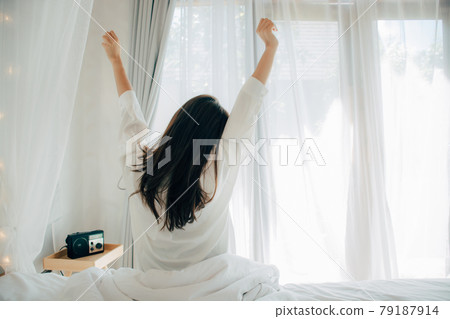 Young asian woman in white pajamas waking up in her bed sitting near the big window fully rested and open the curtains in the morning to get fresh air and sunshine. Happy Young Girl Greets New Day. 79187914