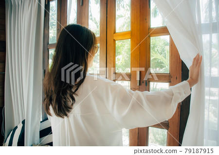 Side view of young woman wearing glasses wake up in bedroom at home or hotel, open white curtains look in window admiring to get fresh air. welcome new day morning feel positive and optimistic.  79187915