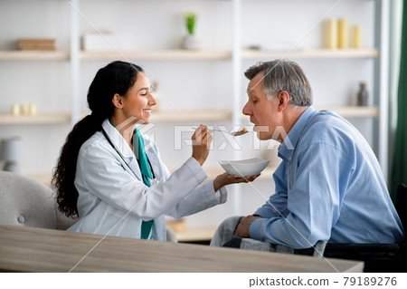 Caregiver feeding elderly man in wheelchair at home. Medical assistance for senior people with partial paralysis Caregiver feeding elderly man in wheelchair at home. Medical assistance for senior people with partial paralysis 79189276