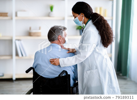 Young female doctor in face mask listening to old man's breathing, using stethoscope at home 79189311