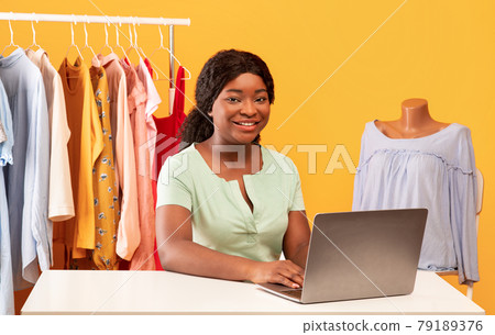 Online shopping. Happy black woman using laptop in showroom, sitting at table near clothes rack, taking web order 79189376