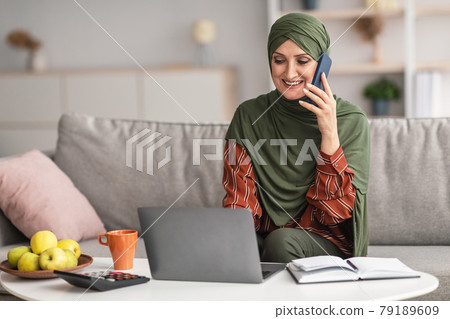 Middle-Aged Islamic Lady Using Laptop Talking On Phone Indoors 79189609