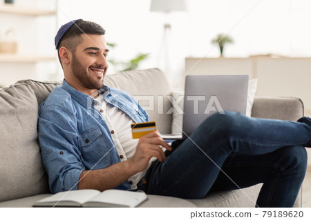 Smiling jewish guy using laptop and credit card at home Smiling jewish guy using laptop and credit card at home 79189620