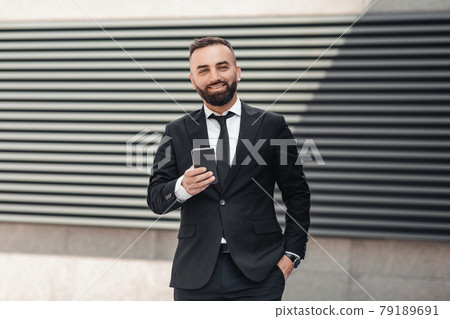 Happy mature entrepreneur in black suit using smartphone, standing near office building with his hand in pocket Happy mature entrepreneur in black suit using smartphone, standing near office building with his hand in pocket 79189691