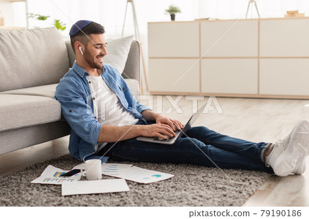 Smiling israeli male working on laptop at home 79190186