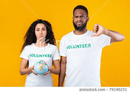 Female And Male Volunteers Holding Globe Gesturing Thumbs-Down, Yellow Background Female And Male Volunteers Holding Globe Gesturing Thumbs-Down, Yellow Background 79190231